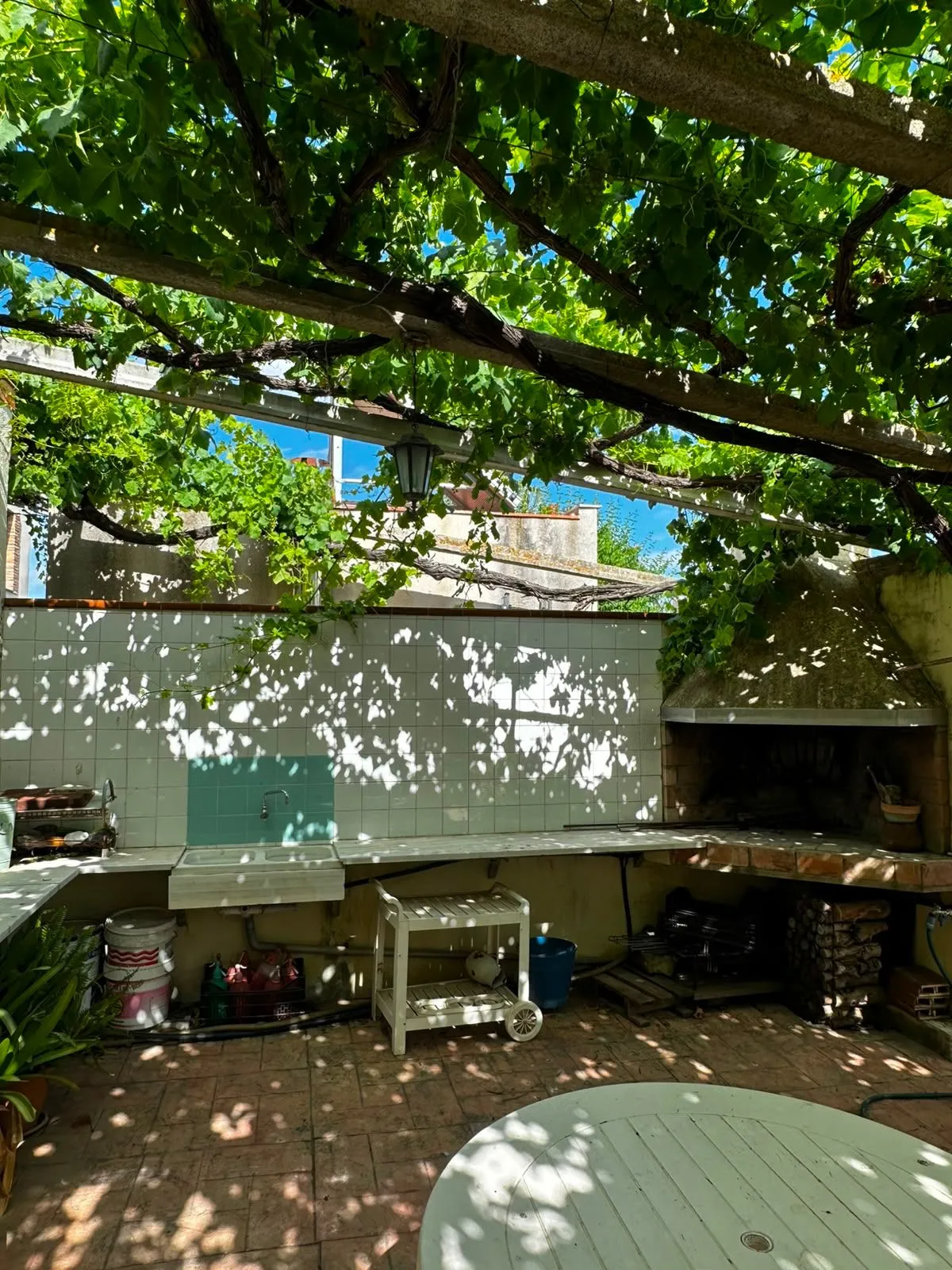 Cocina exterior bajo la parra, con fregadero de obra, encimera de piedra y barbacoa de leña